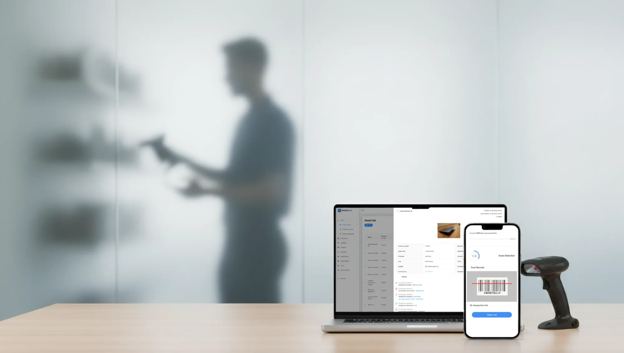 On a desk in front of a frosted glass wall, a laptop shows the AssetLens asset list and one asset's history, a phone scans an asset's barcode during an inventory count, and a handheld barcode scanner stands beside them. Behind the glass, a blurred figure scans items on a shelf.