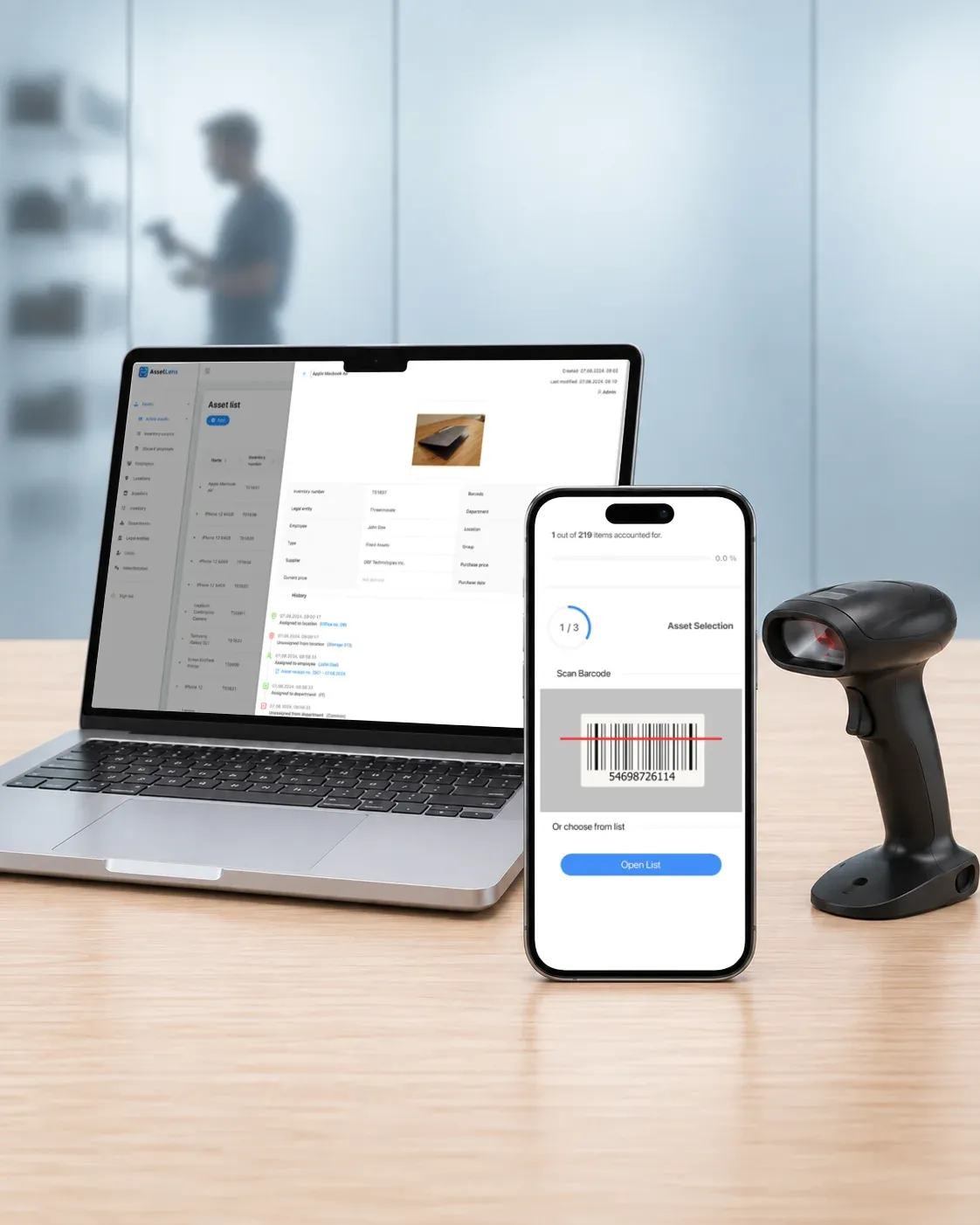 On a desk in front of a frosted glass wall, a laptop shows the AssetLens asset list and an asset's history, a phone scans an asset's barcode during an inventory count, and a handheld barcode scanner stands beside them. Behind the glass, a blurred figure scans items on a shelf.