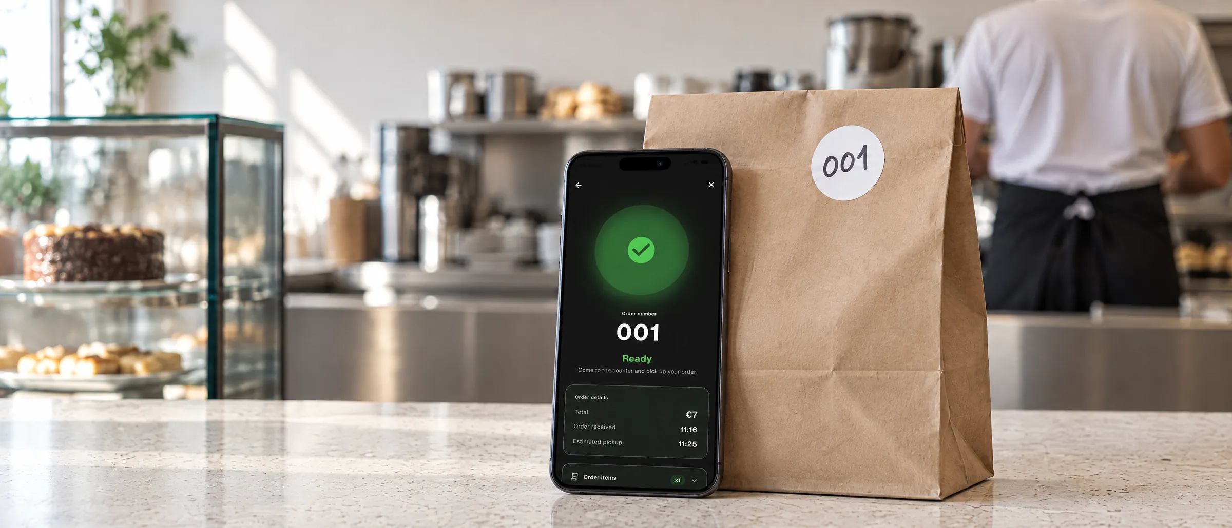 A phone stands on the pickup counter, leaning against a paper takeaway bag with a 001 sticker. The app shows order 001 as ready: come to the counter and pick up your order. Behind, out of focus, the cake display and the kitchen.